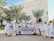 A group of people dressed in white religious attire are standing outdoors in front of a building with decorative architectural features. They are holding a banner that reads 'Umroh Akhir Tahun Talang Mas Tour & Travel' with dates '27 Des 2022 - 04 Jan 2023'. Palm trees flank the group, and everyone appears to be smiling and engaging with the camera.
