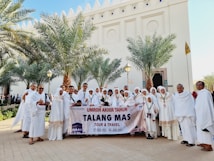 A group of people dressed in white religious attire are standing outdoors in front of a building with decorative architectural features. They are holding a banner that reads 'Umroh Akhir Tahun Talang Mas Tour & Travel' with dates '27 Des 2022 - 04 Jan 2023'. Palm trees flank the group, and everyone appears to be smiling and engaging with the camera.