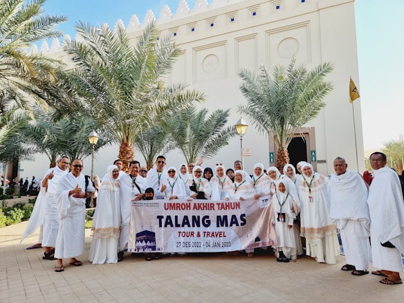 A group of people dressed in white religious attire are standing outdoors in front of a building with decorative architectural features. They are holding a banner that reads 'Umroh Akhir Tahun Talang Mas Tour & Travel' with dates '27 Des 2022 - 04 Jan 2023'. Palm trees flank the group, and everyone appears to be smiling and engaging with the camera.