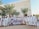 A large group of people in traditional white clothing are gathered in front of a building with palm trees around. They are holding a banner with dates and a travel agency's name. The backdrop includes a tall structure with ornately carved details.