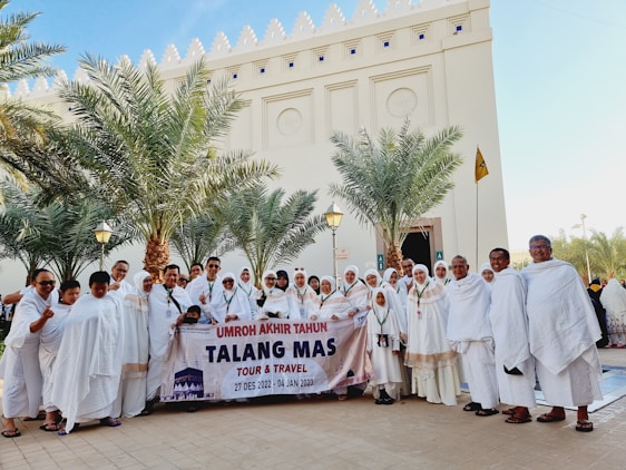 A large group of people in traditional white clothing are gathered in front of a building with palm trees around. They are holding a banner with dates and a travel agency's name. The backdrop includes a tall structure with ornately carved details.