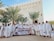 A large group of people in traditional white clothing are gathered in front of a building with palm trees around. They are holding a banner with dates and a travel agency's name. The backdrop includes a tall structure with ornately carved details.
