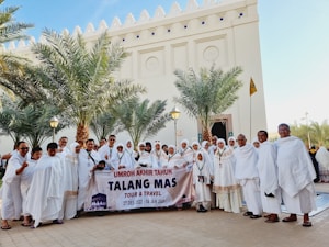 A large group of people in traditional white clothing are gathered in front of a building with palm trees around. They are holding a banner with dates and a travel agency's name. The backdrop includes a tall structure with ornately carved details.