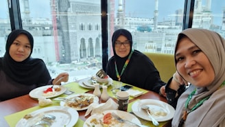 Three women are sitting at a table in a restaurant, enjoying a meal together. They are wearing hijabs and are smiling at the camera. The table is set with plates, cups, and cutlery, and there is food on the plates, such as rice and what appears to be a dessert with watermelon. In the background, a large mosque with intricate architecture can be seen through a large window.