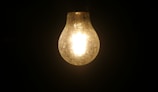A glowing light bulb prominently displayed against a dark background, illuminating its surroundings faintly. The filament inside the bulb is visible due to the bright light it emits, creating a contrast with the darkness around it.