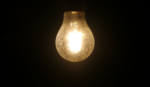 A glowing light bulb prominently displayed against a dark background, illuminating its surroundings faintly. The filament inside the bulb is visible due to the bright light it emits, creating a contrast with the darkness around it.