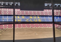 A large football stadium with red and blue seating, featuring the words 'MES QUE UN CLUB' in yellow across the middle section. The pitch is barren, indicating either preparation or maintenance work.