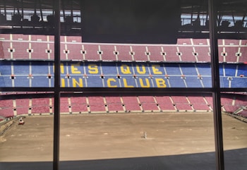 A large football stadium with red and blue seating, featuring the words 'MES QUE UN CLUB' in yellow across the middle section. The pitch is barren, indicating either preparation or maintenance work.