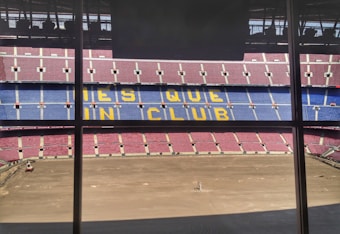A large football stadium with red and blue seating, featuring the words 'MES QUE UN CLUB' in yellow across the middle section. The pitch is barren, indicating either preparation or maintenance work.
