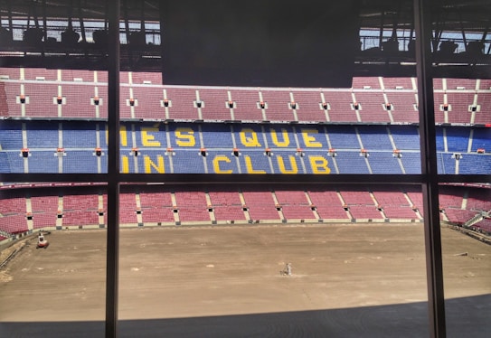 A large football stadium with red and blue seating, featuring the words 'MES QUE UN CLUB' in yellow across the middle section. The pitch is barren, indicating either preparation or maintenance work.