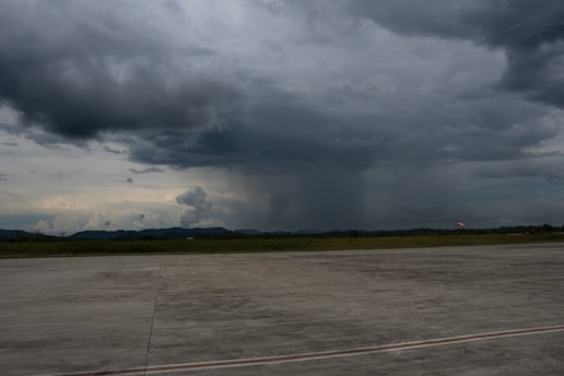 Dark, heavy clouds dominate the sky, suggesting an impending storm. The foreground shows a large concrete surface, likely an airport runway, with green fields and distant hills in the background.