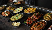 A variety of Indian dishes are arranged on a dark rustic table. The spread includes colorful and flavorful looking food, such as curries, grilled vegetables, and rice, garnished with fresh herbs.