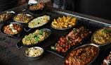 A variety of Indian dishes are arranged on a dark rustic table. The spread includes colorful and flavorful looking food, such as curries, grilled vegetables, and rice, garnished with fresh herbs.