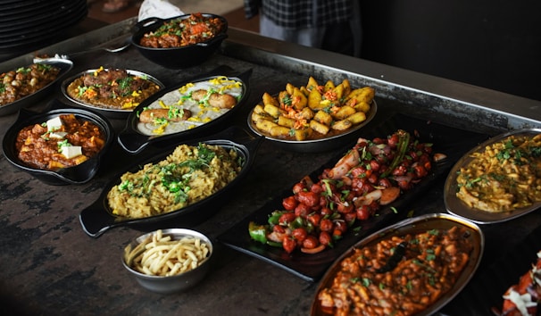 A variety of Indian dishes are arranged on a dark rustic table. The spread includes colorful and flavorful looking food, such as curries, grilled vegetables, and rice, garnished with fresh herbs.