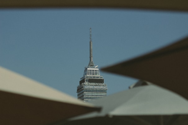 A tall, sharp-tipped skyscraper with the text 'Latino Seguros' is framed by dark shapes in the foreground against a clear blue sky.