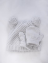 Flat lay of baby accessories including a cotton hat and mittens in white with red details.