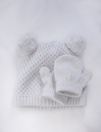 A knitted white hat with two fluffy pom-poms on top, accompanied by a pair of matching mittens, set against a white background. The hat features a textured pattern and ribbed edges.