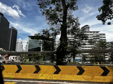 A large urban scene with modern skyscrapers in the background, partially obscured by tall trees. The foreground features a prominent yellow and black road barrier, suggesting a construction or traffic management zone. The sky is clear and blue, with a few scattered clouds overhead.