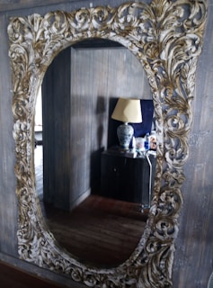 An ornate mirror with an intricate gold and white carved frame hangs on a wall, reflecting a room with wooden paneling. In the reflection, a side table is visible, topped with a decorative lamp featuring a dark floral pattern. Various small objects, including bottles, are also on the table.