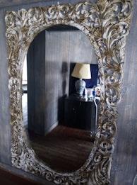 An ornate mirror with an intricate gold and white carved frame hangs on a wall, reflecting a room with wooden paneling. In the reflection, a side table is visible, topped with a decorative lamp featuring a dark floral pattern. Various small objects, including bottles, are also on the table.