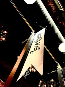 A white decorative banner hangs from the ceiling of an industrial space, featuring black lettering or symbols. The ceiling structure includes visible pipes and fixtures. Overhead lights are on, casting a warm glow and creating a cozy ambiance. The setting appears to be indoors, possibly a restaurant or a public venue with exposed architectural elements.