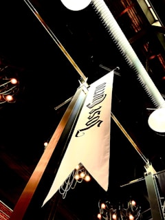 A white decorative banner hangs from the ceiling of an industrial space, featuring black lettering or symbols. The ceiling structure includes visible pipes and fixtures. Overhead lights are on, casting a warm glow and creating a cozy ambiance. The setting appears to be indoors, possibly a restaurant or a public venue with exposed architectural elements.