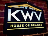 A dark rectangular sign on a brick wall features bold white letters 'KWV' above the words 'House of Brandy' in bright yellow. The sign is bordered by a yellow line on the top, forming a subtle triangular shape resembling a roof, and another line below the text.