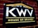 A dark rectangular sign on a brick wall features bold white letters 'KWV' above the words 'House of Brandy' in bright yellow. The sign is bordered by a yellow line on the top, forming a subtle triangular shape resembling a roof, and another line below the text.