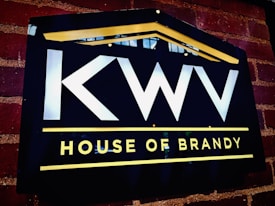 A dark rectangular sign on a brick wall features bold white letters 'KWV' above the words 'House of Brandy' in bright yellow. The sign is bordered by a yellow line on the top, forming a subtle triangular shape resembling a roof, and another line below the text.
