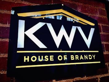 A dark rectangular sign on a brick wall features bold white letters 'KWV' above the words 'House of Brandy' in bright yellow. The sign is bordered by a yellow line on the top, forming a subtle triangular shape resembling a roof, and another line below the text.