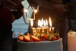 A decorated cake topped with strawberries and gold candles spelling out 'BIRTHDAY' is lit, with sparklers adding a festive touch. In the background, people are gathered, creating a cozy and celebratory atmosphere.