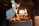 A decorated cake topped with strawberries and gold candles spelling out 'BIRTHDAY' is lit, with sparklers adding a festive touch. In the background, people are gathered, creating a cozy and celebratory atmosphere.