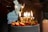 A decorated cake topped with strawberries and gold candles spelling out 'BIRTHDAY' is lit, with sparklers adding a festive touch. In the background, people are gathered, creating a cozy and celebratory atmosphere.
