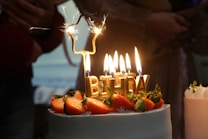 A decorated cake topped with strawberries and gold candles spelling out 'BIRTHDAY' is lit, with sparklers adding a festive touch. In the background, people are gathered, creating a cozy and celebratory atmosphere.
