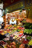 A vibrant marketplace display featuring a wide variety of fresh fruits and vegetables neatly arranged in crates and boxes. There are bundles of garlic, tomatoes, bananas, and other produce hanging above. The setup includes items like peppers, melons, apples, and leafy greens. A vendor stands behind the produce, near a sign that reads 'Alegria de la Huerta'.