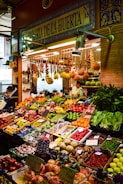 A vibrant marketplace display featuring a wide variety of fresh fruits and vegetables neatly arranged in crates and boxes. There are bundles of garlic, tomatoes, bananas, and other produce hanging above. The setup includes items like peppers, melons, apples, and leafy greens. A vendor stands behind the produce, near a sign that reads 'Alegria de la Huerta'.