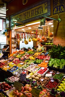 A vibrant marketplace display featuring a wide variety of fresh fruits and vegetables neatly arranged in crates and boxes. There are bundles of garlic, tomatoes, bananas, and other produce hanging above. The setup includes items like peppers, melons, apples, and leafy greens. A vendor stands behind the produce, near a sign that reads 'Alegria de la Huerta'.
