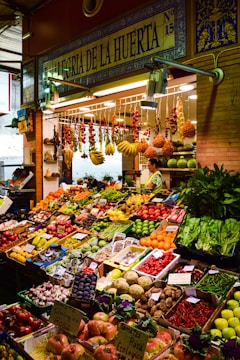 A vibrant marketplace display featuring a wide variety of fresh fruits and vegetables neatly arranged in crates and boxes. There are bundles of garlic, tomatoes, bananas, and other produce hanging above. The setup includes items like peppers, melons, apples, and leafy greens. A vendor stands behind the produce, near a sign that reads 'Alegria de la Huerta'.