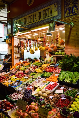 A vibrant marketplace display featuring a wide variety of fresh fruits and vegetables neatly arranged in crates and boxes. There are bundles of garlic, tomatoes, bananas, and other produce hanging above. The setup includes items like peppers, melons, apples, and leafy greens. A vendor stands behind the produce, near a sign that reads 'Alegria de la Huerta'.