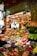 A vibrant marketplace display featuring a wide variety of fresh fruits and vegetables neatly arranged in crates and boxes. There are bundles of garlic, tomatoes, bananas, and other produce hanging above. The setup includes items like peppers, melons, apples, and leafy greens. A vendor stands behind the produce, near a sign that reads 'Alegria de la Huerta'.