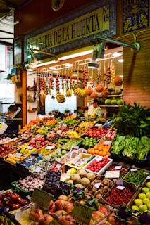 A vibrant marketplace display featuring a wide variety of fresh fruits and vegetables neatly arranged in crates and boxes. There are bundles of garlic, tomatoes, bananas, and other produce hanging above. The setup includes items like peppers, melons, apples, and leafy greens. A vendor stands behind the produce, near a sign that reads 'Alegria de la Huerta'.