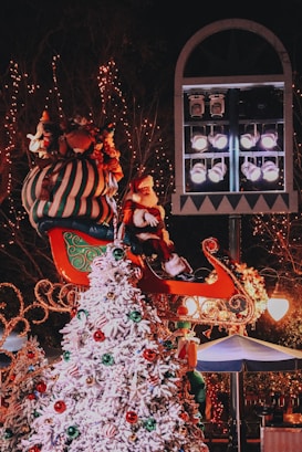 A festive scene features a Christmas display with a Santa figure sitting in a colorful sleigh above a snowy Christmas tree adorned with red, green, and gold ornaments. Surrounding trees and structures are decorated with twinkling lights, and a large spotlight fixture is visible in the background, casting a warm glow over the scene.