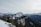 A stunning mountain landscape with snow-covered peaks and dense green forests. A modern building is nestled among the trees on a slope, surrounded by vast mountain ranges under an overcast sky. The scene conveys a sense of stillness and natural beauty.