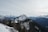 A stunning mountain landscape with snow-covered peaks and dense green forests. A modern building is nestled among the trees on a slope, surrounded by vast mountain ranges under an overcast sky. The scene conveys a sense of stillness and natural beauty.