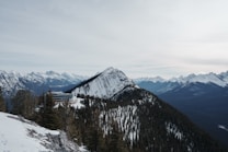 A stunning mountain landscape with snow-covered peaks and dense green forests. A modern building is nestled among the trees on a slope, surrounded by vast mountain ranges under an overcast sky. The scene conveys a sense of stillness and natural beauty.
