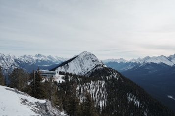 A stunning mountain landscape with snow-covered peaks and dense green forests. A modern building is nestled among the trees on a slope, surrounded by vast mountain ranges under an overcast sky. The scene conveys a sense of stillness and natural beauty.