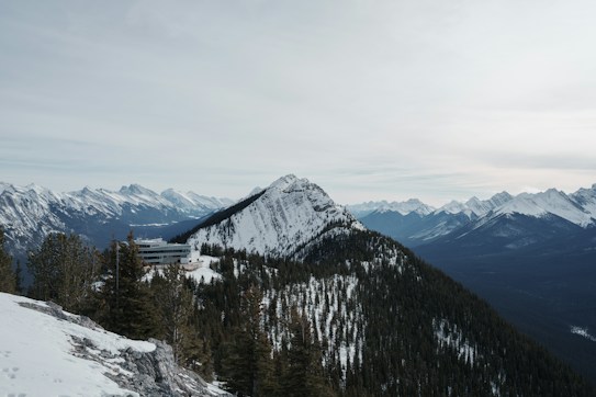 A stunning mountain landscape with snow-covered peaks and dense green forests. A modern building is nestled among the trees on a slope, surrounded by vast mountain ranges under an overcast sky. The scene conveys a sense of stillness and natural beauty.