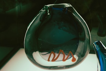 A uniquely shaped glass sculpture with a transparent, almost circular form. The object has a dark blue hue with an orange wavy line on its surface, placed on a light background that enhances its artistic design.