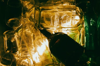 A collection of various transparent glass bottles and jars stacked together with an illuminated background. The light reflects off the glass surfaces, creating a warm glow and highlighting intricate details on the bottles.