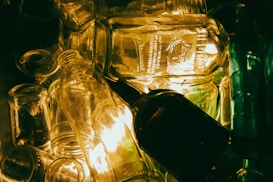 A collection of various transparent glass bottles and jars stacked together with an illuminated background. The light reflects off the glass surfaces, creating a warm glow and highlighting intricate details on the bottles.